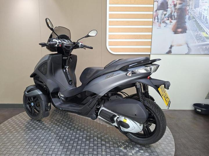 A sleek gray three-wheeled scooter parked on a textured surface, with a modern interior backdrop featuring an abstract display.