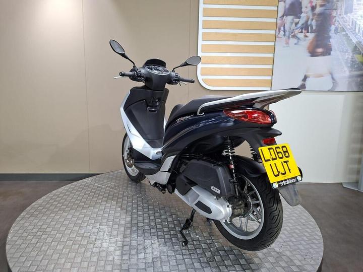 A sleek, blue and white scooter displayed on a circular metal platform, viewed from the rear, with a clear license plate showing "LD68 UJT." The background features a subtle, light-colored wall and a partially visible graphic of people walking.