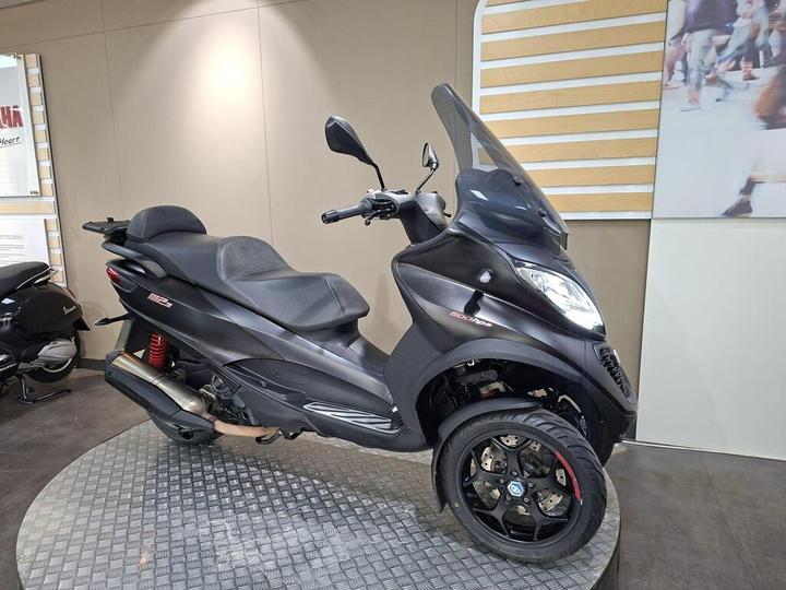 A black three-wheeled scooter displayed on a circular platform in a showroom, featuring a large windshield and sporty design, with red suspension visible and a sleek black finish.