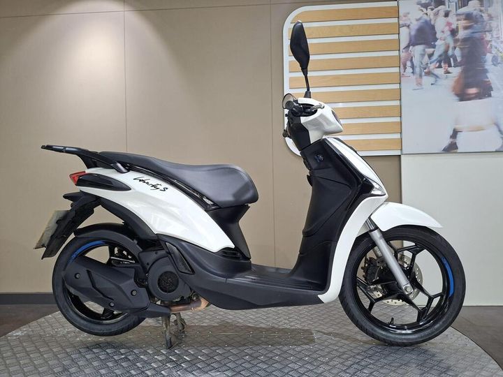 Piaggio Liberty S 125 (2025) A side view of a white scooter with black accents, featuring a sleek design, visible wheels, and a modern seat, displayed on a textured surface against a neutral background.
