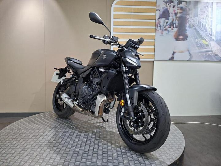 A black motorcycle displayed on a circular platform in a minimalist showroom setting, featuring a clean design with prominent tires, visible engine components, and sleek handlebars. In the background, there is a large photograph of people walking in an urban environment.