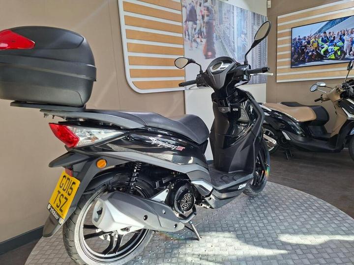 A sleek black scooter with a rear top case, positioned against a stylish interior backdrop featuring wood paneling and wall photos, with another scooter partially visible on the right.