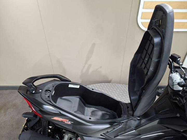 Close-up of a black scooter with an open seat, revealing a storage compartment underneath. The background features a light-colored wall.