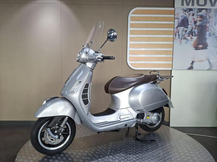 A silver Vespa scooter displayed on a circular platform against a neutral wall, featuring a clear windscreen and a brown seat.