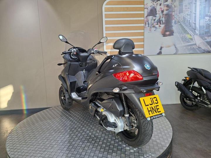 A gray three-wheeled scooter displayed on a circular platform against a light-colored wall, with a blurred cityscape in the background. The scooter features a prominent rear light and a license plate visible at the back.