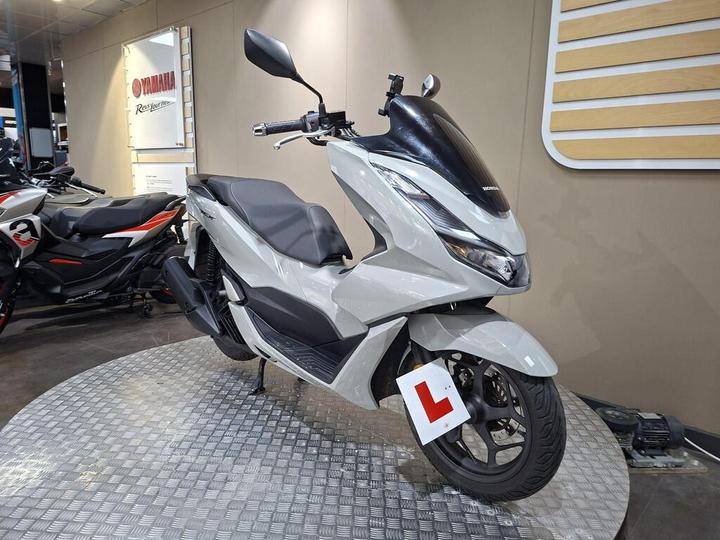 A sleek, modern white scooter with a prominent front shield, displayed on a checkered platform in a Yamaha showroom, featuring a learner plate on the front.