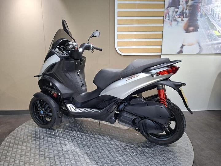 A modern three-wheeled scooter, featuring a sleek white and black design, is showcased on a textured circular platform indoors, with a neutral wall backdrop and a large image of pedestrians in a cityscape visible on the right.