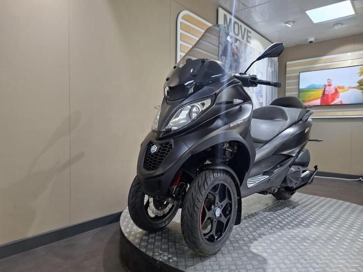 A black three-wheeled scooter displayed on a circular platform in an indoor showroom setting, featuring a windshield and sleek design.