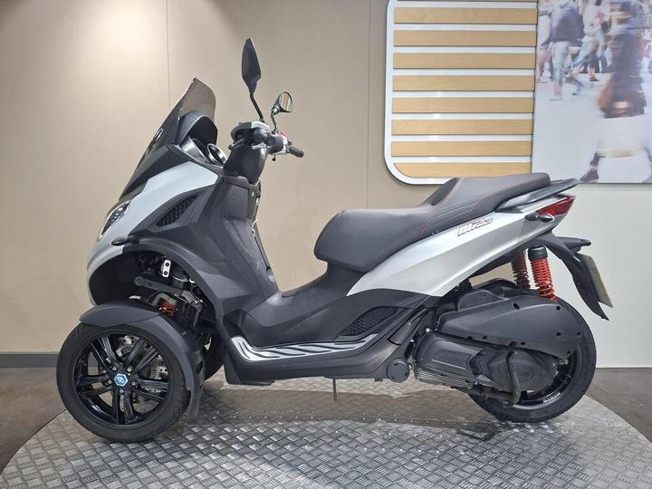 A sleek, silver and black three-wheeled scooter parked on a textured floor, showcasing its modern design and features.