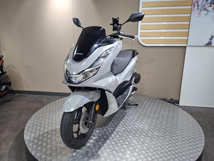 A silver Honda scooter displayed on a circular platform, featuring a sleek design and modern details, set against a neutral background.
