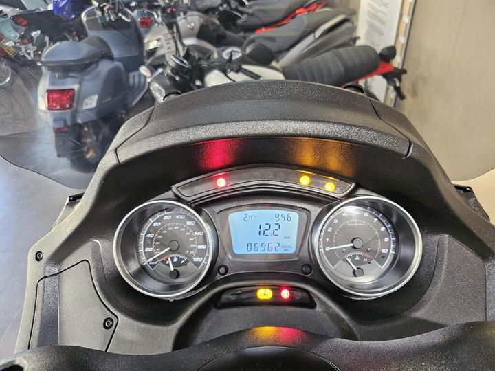 Close-up view of a motorcycle dashboard displaying digital and analog gauges, including speedometer, odometer showing 06962 miles, and indicator lights. Background features other motorcycles and scooters.