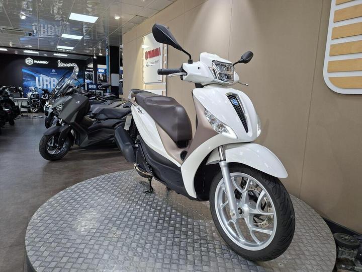 A white scooter with a brown seat is displayed on a circular platform in a motorcycle showroom, with a dimly lit background featuring other bikes and promotional signage.