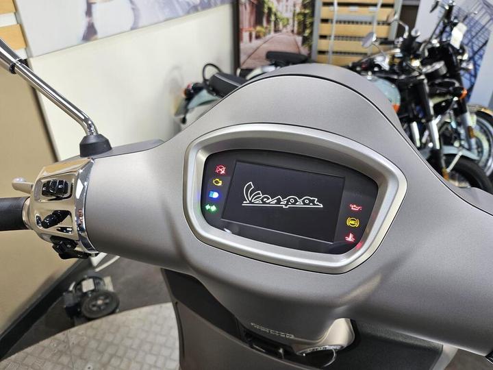 Close-up view of a scooter's dashboard featuring a digital display with the "Vespa" logo, various indicator lights, and chrome control switches, set in a showroom with other motorcycles visible in the background.