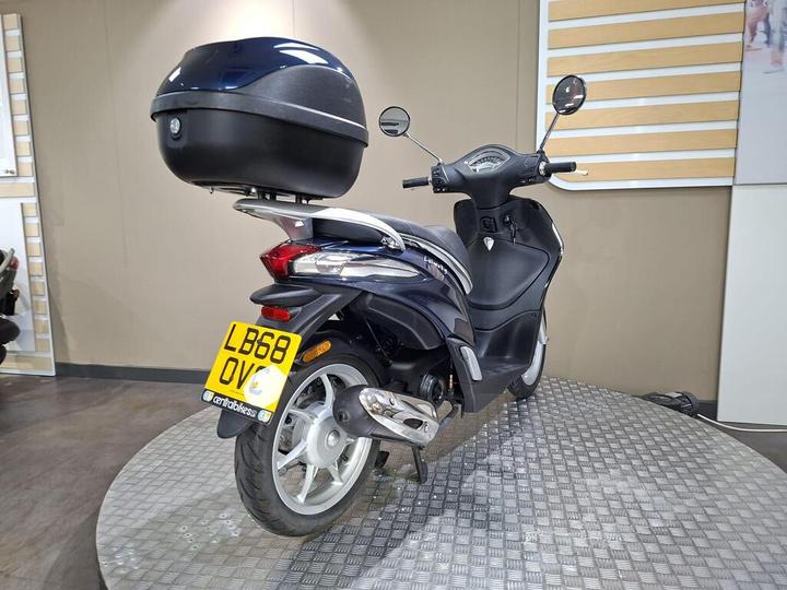 A dark blue scooter with a top case stands on a display platform, showcasing its rear view. The license plate is visible, displaying "LB68 OQV." The background features a neutral-toned wall and a textured floor.