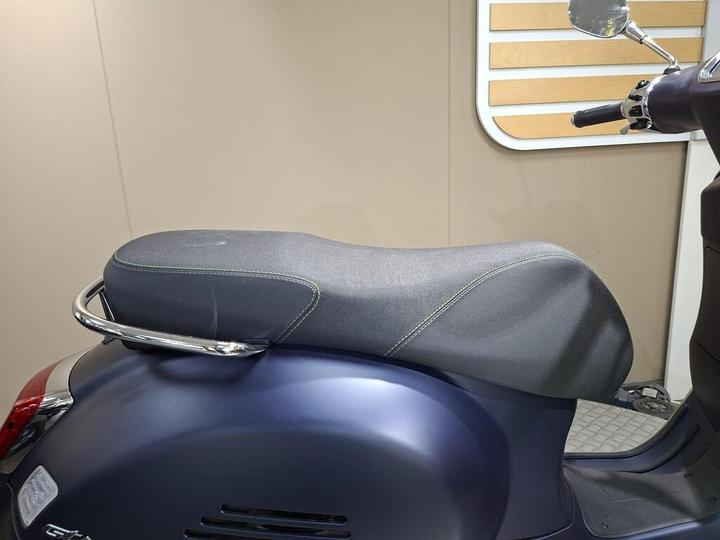 A close-up view of a scooter seat with a sleek design, featuring a dark fabric cover and chrome accents, set against a light-colored background.