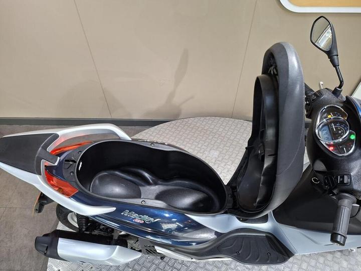 A top-down view of a scooter with an open seat revealing a storage compartment underneath, set on a patterned floor with a plain wall background.