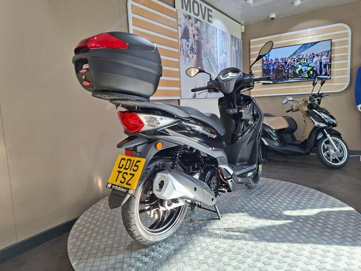 A black scooter with a rear luggage box is parked on a textured surface, set against a modern display featuring a television and light wood accents. A second scooter is visible in the background.