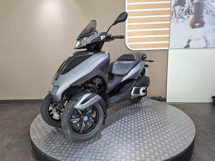A gray three-wheeled scooter displayed on a circular platform, featuring a sleek design, tinted windshield, and a textured front panel, set against a neutral background with a wooden accent wall.