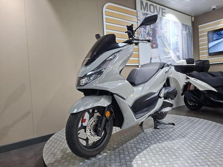 A modern gray scooter displayed on a metal platform in a showroom, with a sleek design, visible front wheel, and a digital screen in the background showcasing urban imagery.