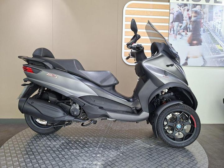 Piaggio MP3 350 LT Sport (2019) A side view of a gray three-wheeled scooter displayed on a textured floor, with a neutral background featuring a branding element and blurred people.