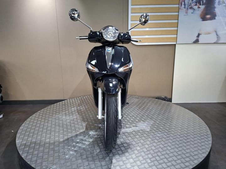 A sleek black scooter positioned frontally on a circular display platform, showcasing its headlights, mirrors, and tires in a modern showroom setting.