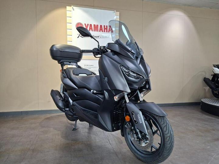 A sleek, matte black Yamaha scooter is displayed in a showroom, featuring a dual seat, a windscreen, and a top box on the rear. A Yamaha banner hangs in the background against a neutral wall.