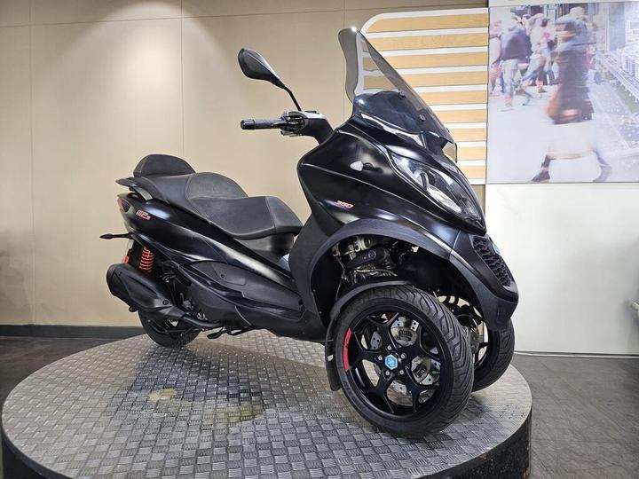 A black three-wheeled scooter parked on a round display platform, showcasing its sleek design and prominent front windshield. The background features a light-colored wall.