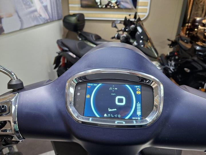 A close-up view of a scooter's digital dashboard displaying digital speed (0 mph), trip distance (1 mile), temperature (19°C), and clock (10:01 AM) with control buttons and motorcycle silhouettes in the background.