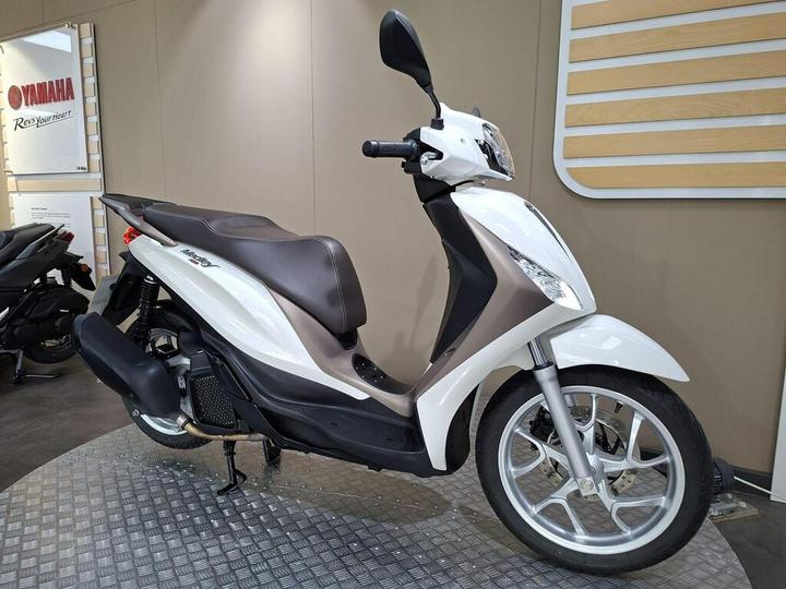 A white and silver scooter displayed in a showroom, featuring a sleek design, a comfortable seat, and positioned on a patterned floor.