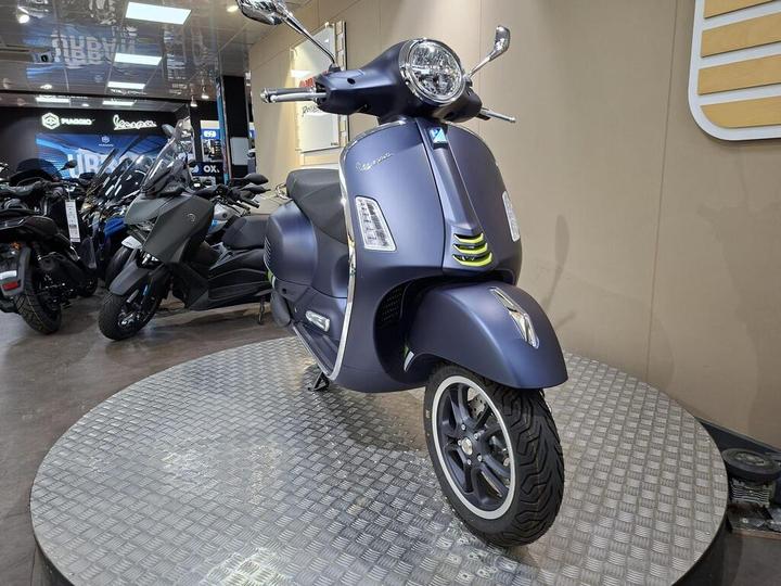 A blue-gray scooter displayed on a circular metal platform within a motorcycle showroom, surrounded by various motorcycles in the background.
