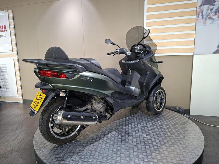 A green three-wheeled scooter displayed on a circular platform, featuring a windshield and a sleek design, with a registration plate visible at the rear.