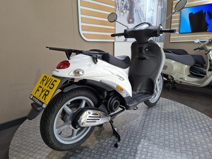 A white Vespa scooter parked on a silver diamond-patterned floor, with a visible license plate reading "RV15 FYR," set against a minimalist interior featuring wooden accents and a blurred background of another scooter display.