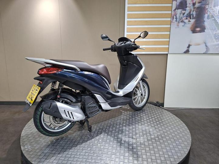 A blue and white scooter is displayed on a circular, textured platform against a neutral background, with a partial image of a busy street visible in the backdrop.