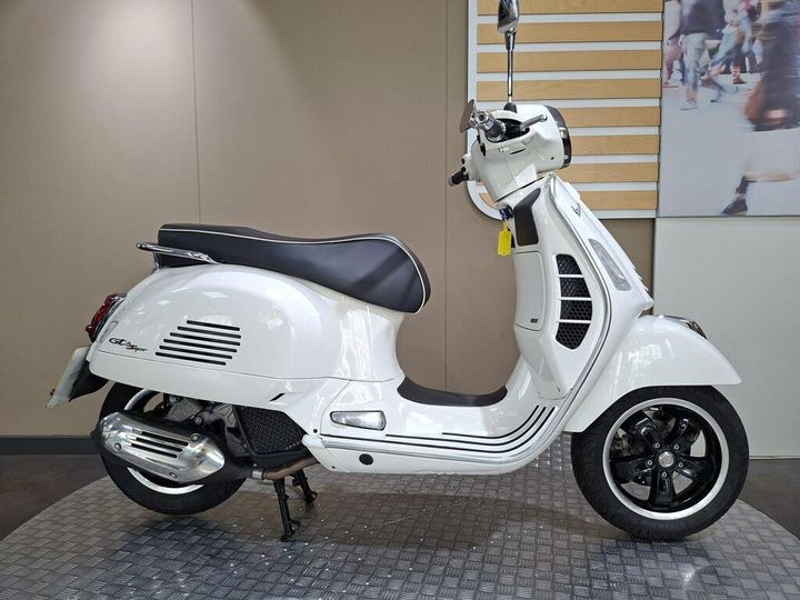 Vespa GTS 125 (2021) A stylish white scooter with a black seat, chrome details, and unique design features, positioned against a neutral background in a showroom setting.