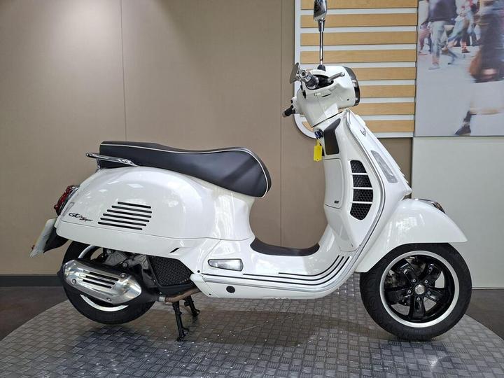 Vespa GTS 125 (2021) A stylish white scooter with a black seat, chrome details, and unique design features, positioned against a neutral background in a showroom setting.