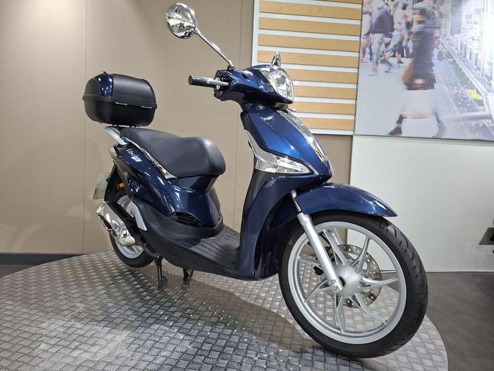 A blue scooter is displayed on a textured platform, featuring a comfortable seat and a black storage compartment on the back, with a backdrop of a blurred urban setting.