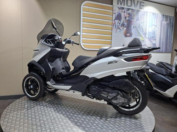A sleek white and black three-wheeled scooter displayed on a rotating platform in a modern showroom, with a backdrop featuring cityscape imagery.