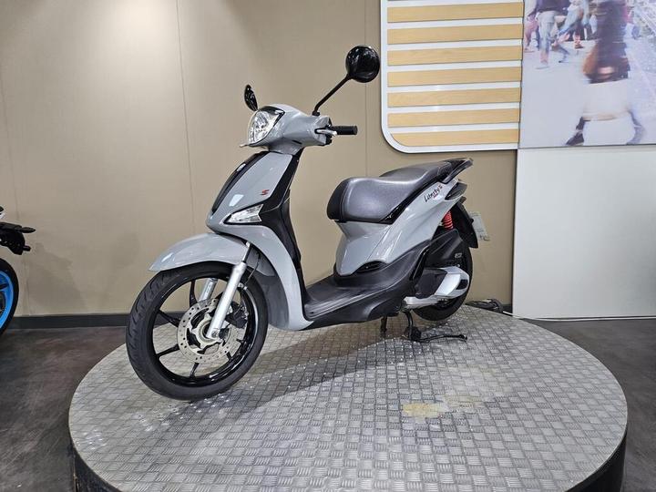 A gray scooter displayed on a circular platform, featuring a modern design, front disc brake, and a cushioned seat. The backdrop shows a neutral-colored wall and a blurred image of people walking.