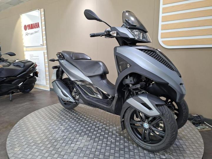 A stylish gray scooter with three wheels displayed on a circular metal platform, set against a neutral background featuring a Yamaha sign.