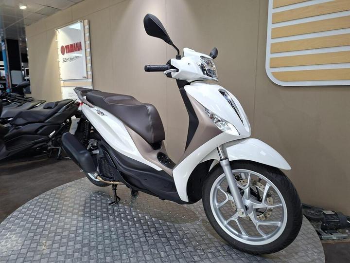 A white and silver scooter displayed on a metal platform, with a blurred background featuring additional scooters and a Yamaha sign.