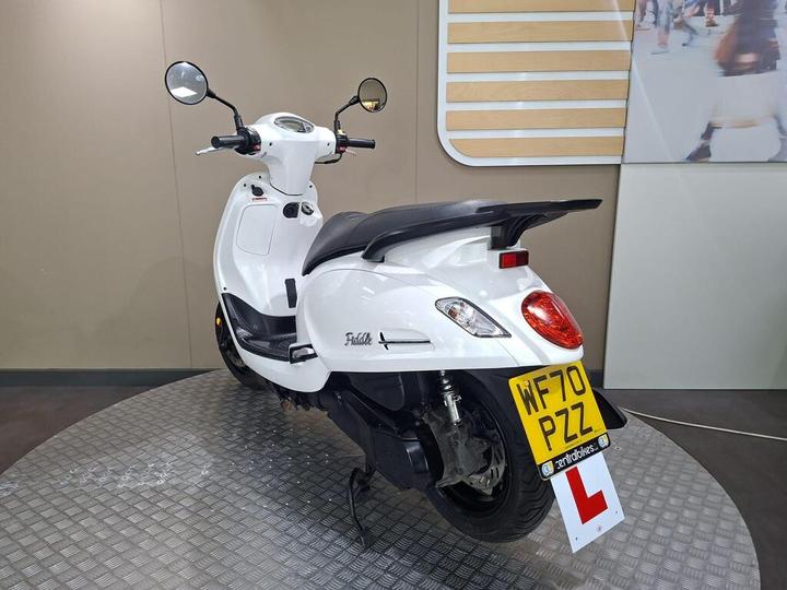 A white scooter parked on a circular display, featuring a learner plate, with a smooth design and dual mirrors. The background includes a simple wall.