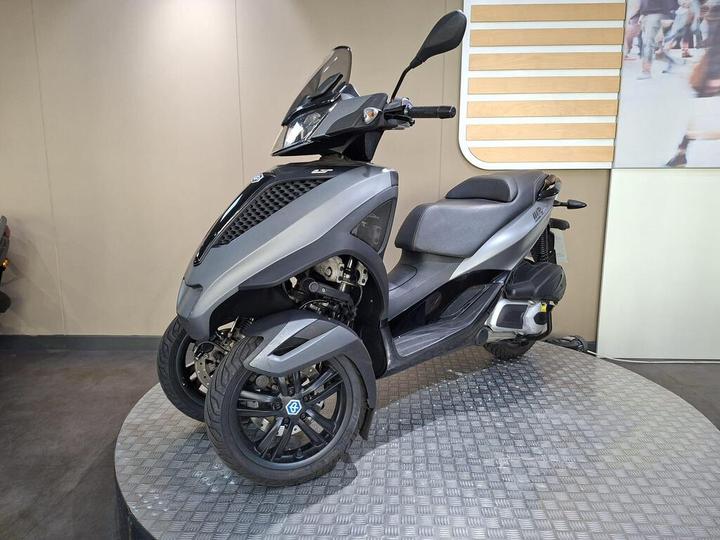 A sleek, modern three-wheeled scooter stands on a circular display pedestal, showcasing its gray body, sporty design, and large front windscreen. The background features a simple, light-colored wall with horizontal wooden slats.