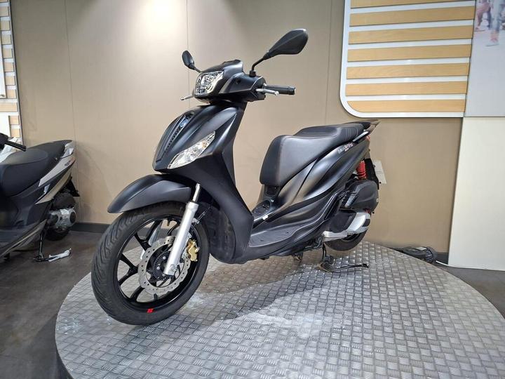 A black scooter is displayed on a circular platform in a modern showroom, with a minimalist background featuring wooden paneling. The scooter has a sleek design with a prominent front headlight and silver accents on the front wheel and brakes.