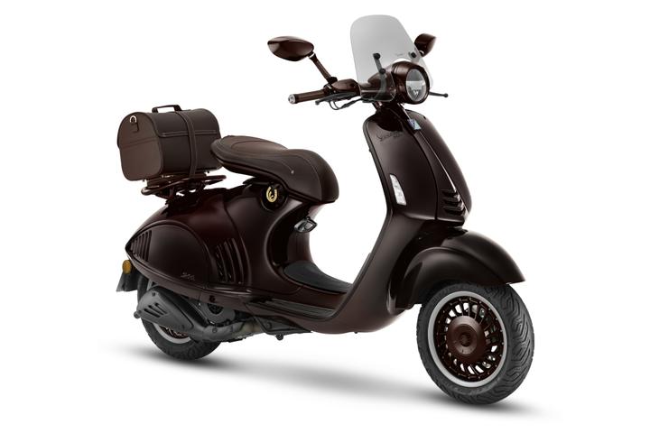 A dark brown vintage-style scooter with a windscreen, a stylish padded seat, and a matching brown saddlebag attached to the rear. The scooter features intricate design details and a distinctive wheel pattern.