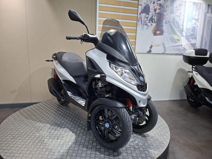 A modern three-wheeled scooter with a sleek silver and black design, showcased on a circular platform with a textured surface, against a neutral background featuring a cityscape mural.