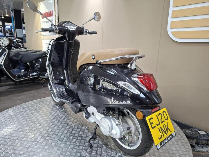 A black Vespa scooter with a tan seat is displayed in a showroom, showcasing its sleek design and chrome accents. The background features other motorcycles and a promotional Yamaha sign. The scooter’s license plate reads "EJ20 JWK."
