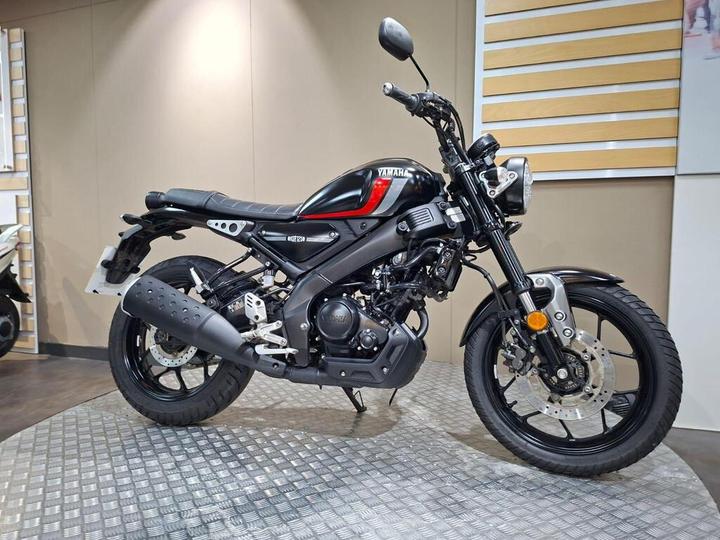 A black Yamaha motorcycle with a red stripe is displayed on a textured platform in a showroom, with a neutral background showcasing the bike's design and features.