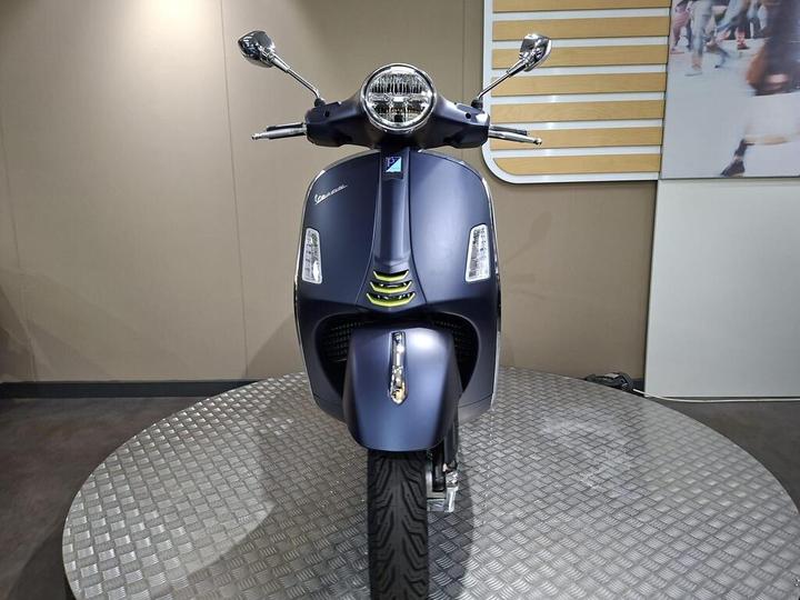 A front view of a blue Vespa scooter, featuring sleek lines, chrome accents, and a round headlight, displayed on a circular metal platform against a neutral background.