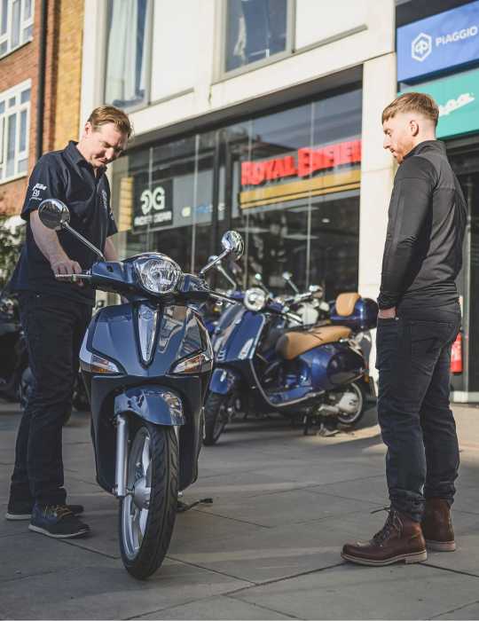 A male motorcycle mechanic inspecting a blue scooter while a customer watches