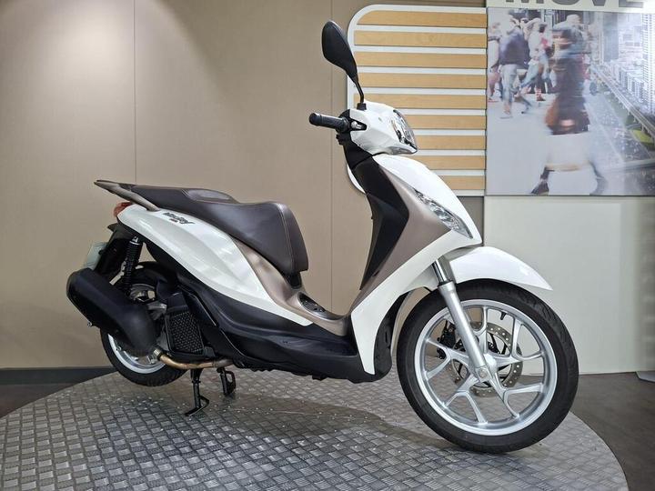 A white and bronze scooter displayed on a textured surface, set against a neutral background with a blurred image of pedestrians.